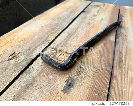Close up view of steel pry bar placed diagonally on rough wooden workbench, captured in natural light, highlighting texture, wear marks and rustic workshop atmosphere Close up view of steel pry bar placed diagonally on rough wooden workbench, captured in natural light, highlighting texture, wear marks and rustic workshop atmosphere 127478298