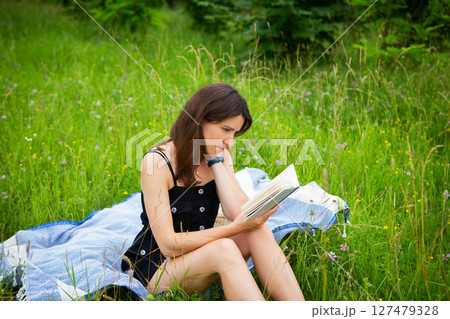 A young woman sits in a grassy field, engrossed in a book, enjoying a peaceful moment outdoors on a summer day 127479328
