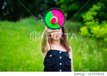 A young woman playfully holds a vibrant, two-toned disc with a neon ball in front of her face in a lush green field 127479329