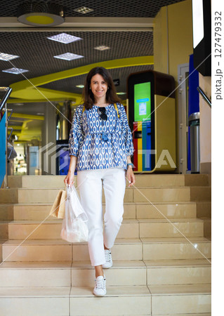 A smiling woman with shopping bags descends stairs in a modern shopping mall, showcasing casual summer fashion A smiling woman with shopping bags descends stairs in a modern shopping mall, showcasing casual summer fashion 127479332