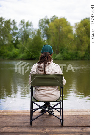 A woman sits on a portable chair by a calm river, enjoying the serene natural landscape 127479341