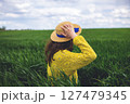 A woman in a yellow sweater and straw hat enjoys a sunny day in a vibrant green field 127479345