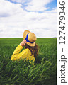 A woman in a yellow sweater and straw hat enjoys the tranquility of a green field, lost in contemplation under a partly cloudy sky 127479346