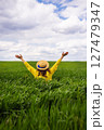 A woman in a yellow sweater and straw hat joyfully embraces a lush green field, arms outstretched under a partly cloudy sky 127479347