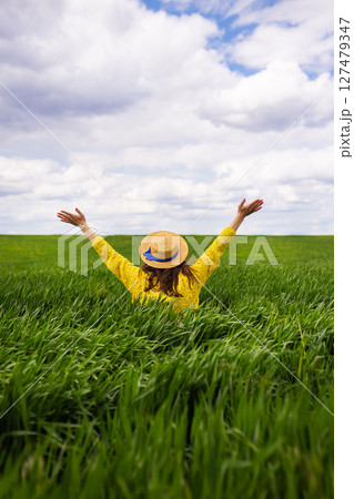 A woman in a yellow sweater and straw hat joyfully embraces a lush green field, arms outstretched under a partly cloudy sky A woman in a yellow sweater and straw hat joyfully embraces a lush green field, arms outstretched under a partly cloudy sky 127479347