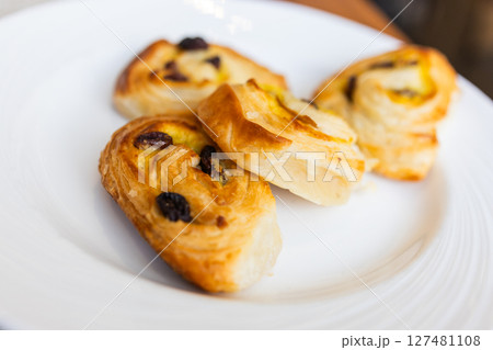 Three raisin pastries served on white plate during hotel breakfast. Morning dessert, sweet bakery snack, and elegant table setting concept. Three raisin pastries served on white plate during hotel breakfast. Morning dessert, sweet bakery snack, and elegant table setting concept. 127481108