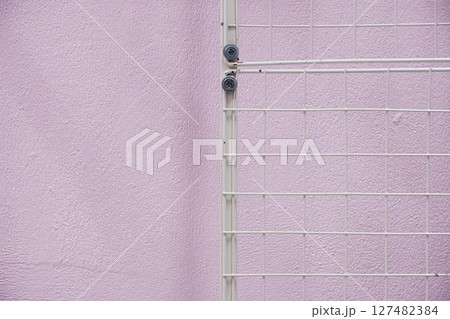 White metal grid fence against a textured pastel pink wall 127482384
