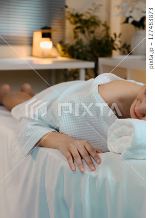 Peaceful spa scene with woman in white robe resting on soft bed preparing for massage 127483873