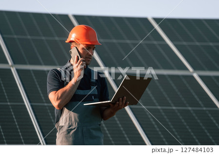 Talking by phone, holding laptop. Man is doing operating and maintenance in solar power plant Talking by phone, holding laptop. Man is doing operating and maintenance in solar power plant 127484103