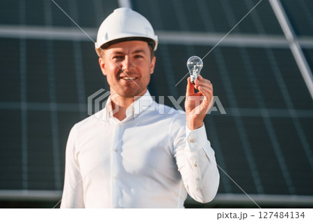 Man in white shirt and hart hat is holding bulb on a solar power plant 127484134