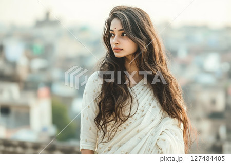Elegant Indian woman with wavy hair in white saree, cityscape backdrop blurred, calm design. 127484405