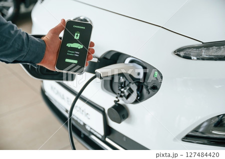 Using smartphone app for monitoring charging process. Close up view of man that is with electrical car Using smartphone app for monitoring charging process. Close up view of man that is with electrical car 127484420