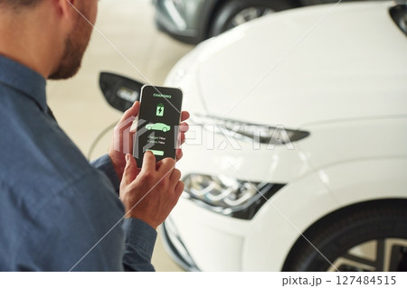 App interface of charging in smartphone. Handsome car dealership worker is with electric vehicle 127484515