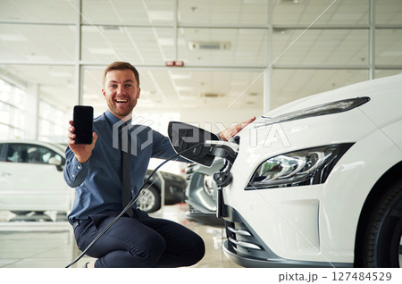 Sitting, showing smartphone, charging. Handsome car dealership worker is with electric vehicle Sitting, showing smartphone, charging. Handsome car dealership worker is with electric vehicle 127484529