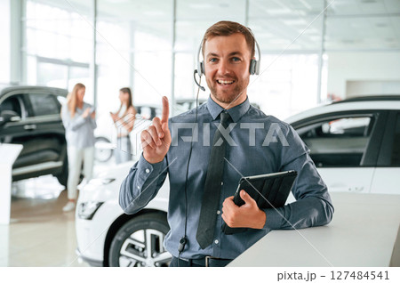 Manager is in the headphones and with notepad. Handsome car dealership worker is with electric vehicle 127484541