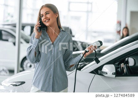 Talking by phone and holding charger. Beautiful woman is indoors with the modern car Talking by phone and holding charger. Beautiful woman is indoors with the modern car 127484589