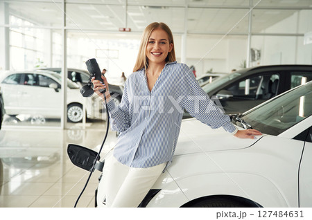 Leaning on the vehicle and holding charger for electric car. Beautiful woman is indoors 127484631