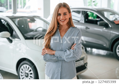 Standing with arms crossed. Beautiful woman is indoors with the modern car Standing with arms crossed. Beautiful woman is indoors with the modern car 127485399