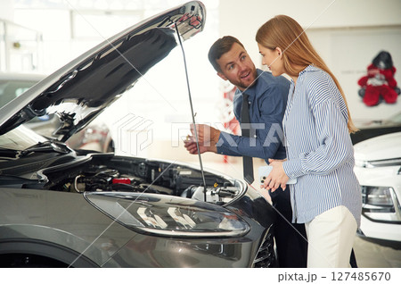 Under the hood. Man in formal clothes is with woman customer with the electric car Under the hood. Man in formal clothes is with woman customer with the electric car 127485670