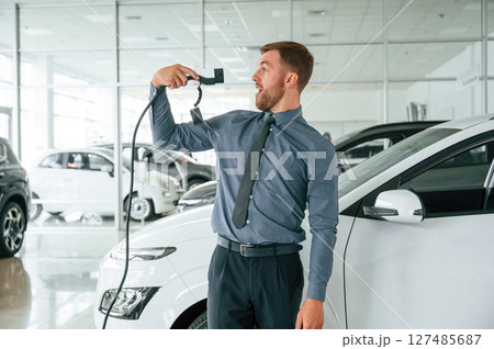 Smiling, holding the charger. Handsome car dealership worker is with electric vehicle Smiling, holding the charger. Handsome car dealership worker is with electric vehicle 127485687