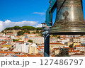 Close-up of historic bell in selective focus overlooking Lisbon, Portugal, colorful rooftops, iconic St Jorge castle, clear blue sky and some clouds in blurred background. City heritage, vibrant 127486797