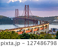 Iconic 25 de Abril bridge in Lisbon, Portugal, in heavy traffic, full with vehicles in transit at sunset. Red suspension bridge over Tagus River, major city landmark, transport route, lush greenery 127486798