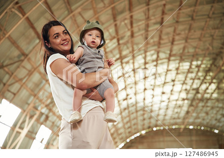 Holding kid in hands. Gorgeous woman with her little baby son is with horse indoors 127486945