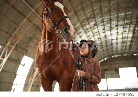 In jockey clothes. Cute little girl is with horse indoors 127487059