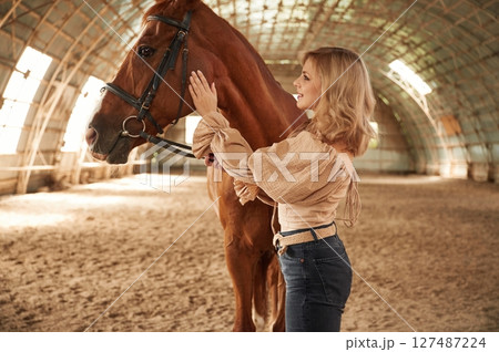In jeans and creamy colored clothes. Beautiful young woman is with horse indoors 127487224
