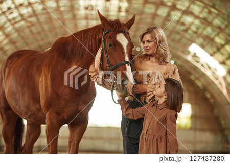 Woman with her little daughter is with horse indoors Woman with her little daughter is with horse indoors 127487280