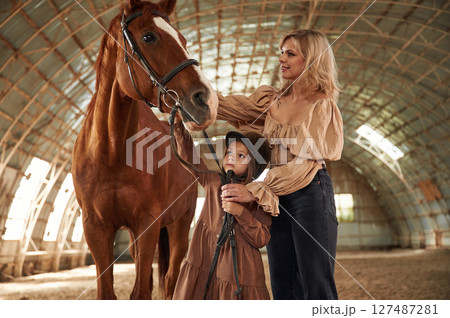 Woman with her little daughter is with horse indoors Woman with her little daughter is with horse indoors 127487281