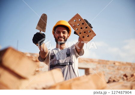 Showing and holding little shovel and brick. Handsome Indian man is on the construction site 127487904