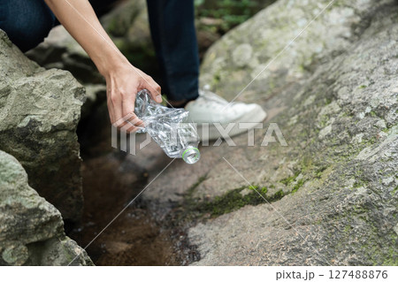 Individual Picking Up Plastic Bottle from Rocks. Volunteer actively engaging in environmental cleanup by removing litter. 127488876