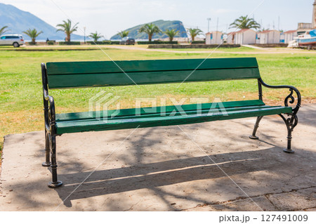 Green wooden bench in sunny coastal park. Public space, relaxation area, and outdoor urban furniture near the sea. 127491009