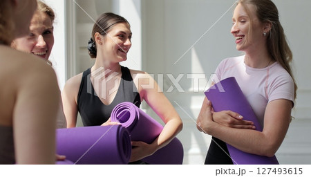 Young women chatting standing with mats in hands after group training in yoga studio. 127493615
