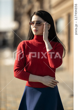 fashionable girl wearing glasses stands confidently on sunny city street She sports cozy red sweater and chic blue skirt while posing gracefully amidst elegant architecture fashionable girl wearing glasses stands confidently on sunny city street She sports cozy red sweater and chic blue skirt while posing gracefully amidst elegant architecture 127494856