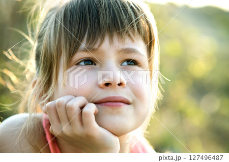 Portrait of pretty child girl with gray eyes and long fair hair leaning on her hands smiling happily outdoors on blurred bright background. Cute female kid on warm summer day outside. 127496487
