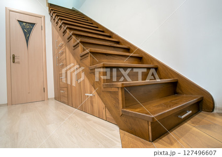 Modern architecture interior with luxury hallway with glossy wooden stairs in multi-storey house. Custom built pullout cabinets on glides in slots under stairs. Use of space for storage. 127496607