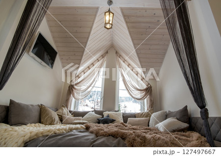 Interior of a modern house spacious hallway with big soft resting place. Contemporary wide sofa with many pillows and light window onder wooden loft ceiling. Interior of a modern house spacious hallway with big soft resting place. Contemporary wide sofa with many pillows and light window onder wooden loft ceiling. 127497667