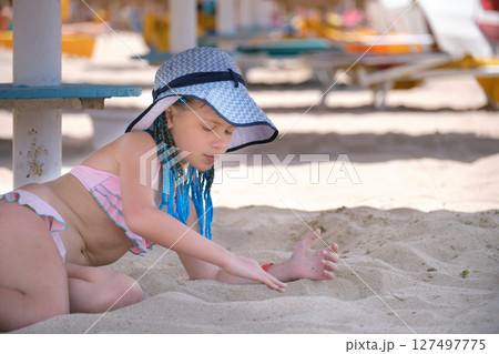 Happy child girl in swimsuit playing with sand under umbrella shadow during summer tropical vacations 127497775