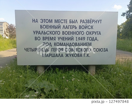 A sign in Russian detailing the history of a military camp established in 1949 under the command of a Soviet hero. Surrounded by greenery and grass. 127497848