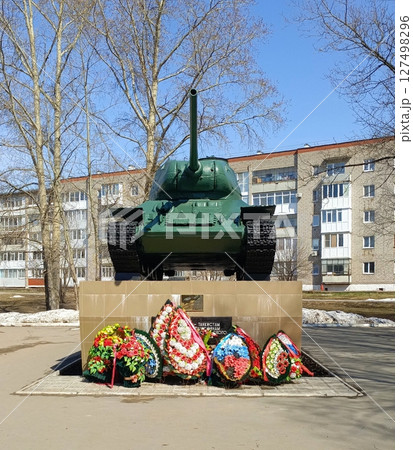 A green tank monument stands on a stone pedestal. Colorful floral wreaths are placed at its base. Surrounding buildings are visible in the background. A green tank monument stands on a stone pedestal. Colorful floral wreaths are placed at its base. Surrounding buildings are visible in the background. 127498296