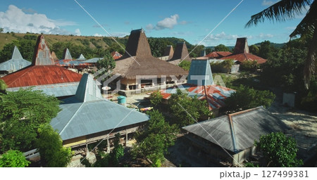 Aerial view of a village in Sumba, Indonesia, showing traditional houses with conical roofs made of straw and metal sheets, surrounded by lush vegetation under a clear blue sky 127499381