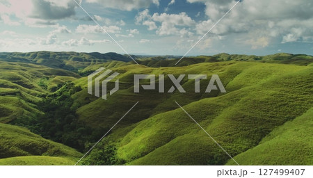Aerial view of the undulating landscape of Sumba Island, Indonesia, showcasing rolling green hills stretching as far as the eye can see under a dramatic sky with fluffy white clouds 127499407