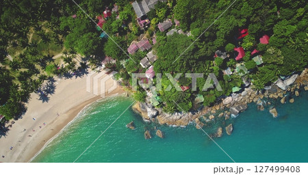 Turquoise water gently wash sand beach and rocky coast, lush tropical vegetation and charming bungalows in Thailand idyllic paradise. Exotic travel vacation getaway. Drone flight. Aerial top down view 127499408