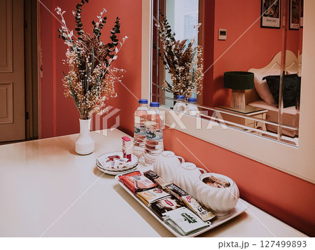 Boutique hotel interior with mirror, flowers, and tray of tea, snacks, and bottled water styled on a clean vanity in a soft coral-colored guest room 127499893