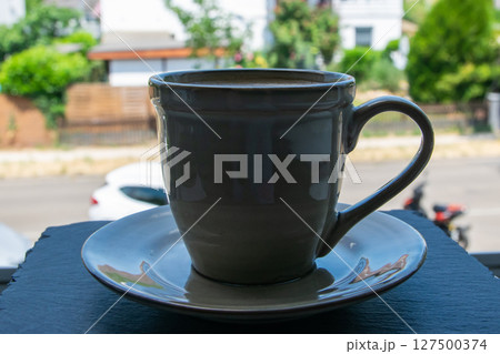 Green cup of coffee stay on the black stone window sill with a beautiful view of the summer street.  127500374
