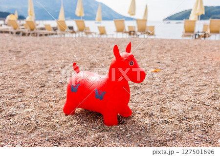 Red inflatable toy on a pebble beach with sun loungers and sea view. Childhood joy, summer vacation, and beachside relaxation. 127501696
