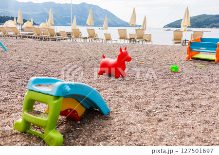 Colorful beach toys scattered on a pebbled shore near sun loungers and sea. Childhood freedom, summer leisure, and playful atmosphere. 127501697
