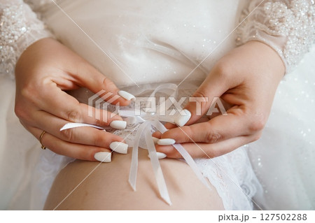 Close-up of Bride's Hands Tying White Ribbon, Wedding Garter Detail, Delicate Lace, Pearl Accents, Soft Focus, Romantic Bridal Scene. Close-up of Bride's Hands Tying White Ribbon, Wedding Garter Detail, Delicate Lace, Pearl Accents, Soft Focus, Romantic Bridal Scene. 127502288